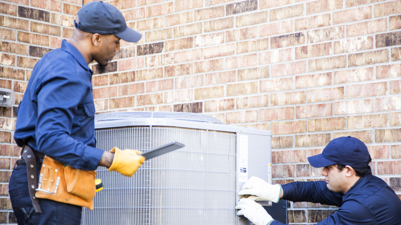 Two people wearing blue uniforms inspecting an AC unit outdoors in front of a brick house