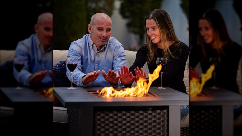 A smiling couple warm their hands in front of a roaring fire pit