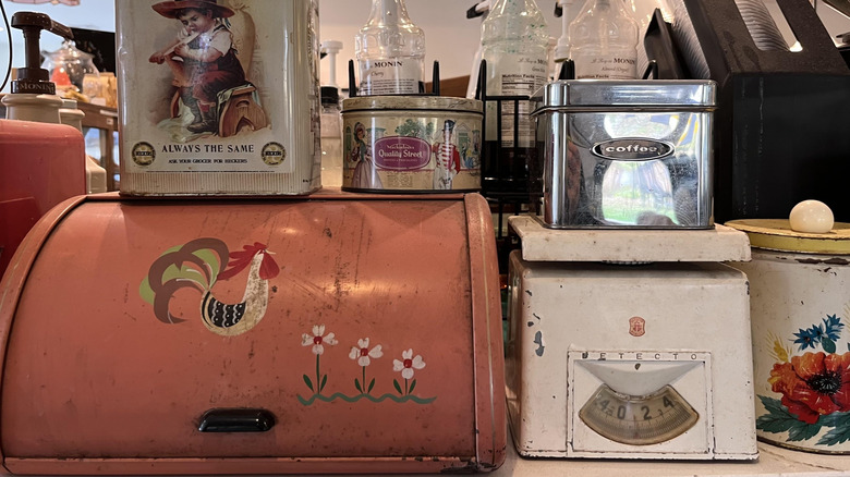 A vintage pink breadbox is displayed at a thrift store surrounded by metal tins
