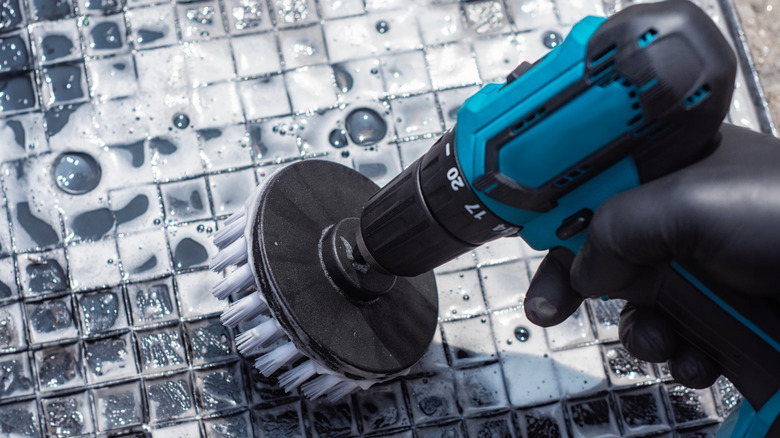 Person using a drill with a cleaning brush attachment to scrub a dark colored floor mat