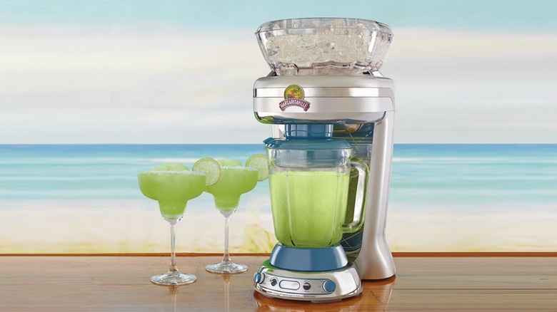 A margarita machine filled with green drink sits next to two full margarita glasses of the same green drink