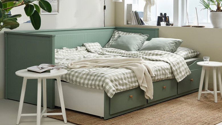 A sage green HEMNES Daybed frame with 3 drawers flanked by two white stools sits in front of a large window