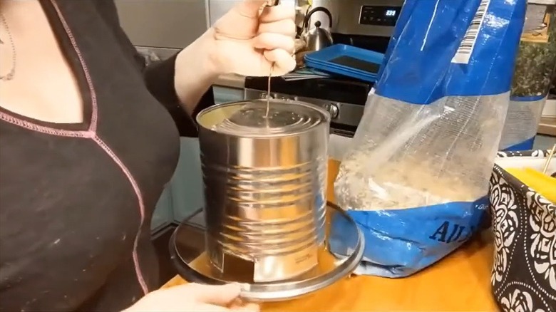 Woman holding DIY bird feeder made from soup can and pot lid in kitchen above wood countertop