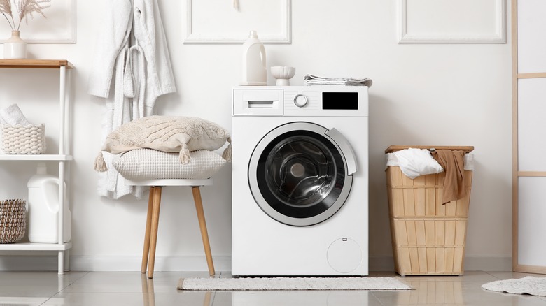A modern washing machine in a bright white laundry room with pillows and laundry basket.