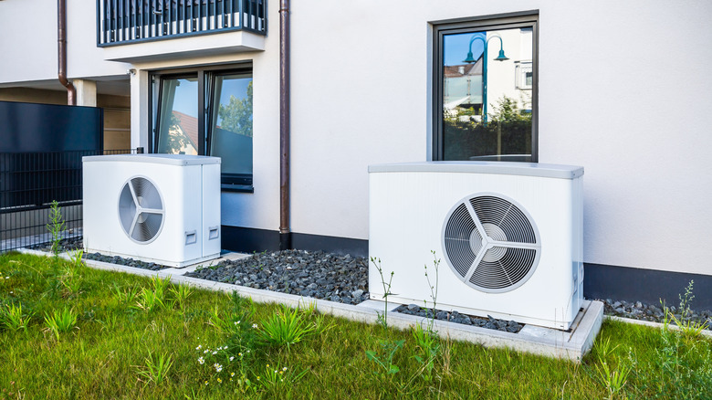 Two energy efficient heat pumps installed outside of a modern multi-unit residential building, on a gravel pad flanked by green grass.