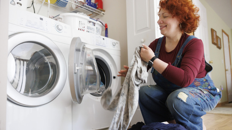 A smiling woman crouching to take laundry out of a dryer, with a matching washing machine next to it.