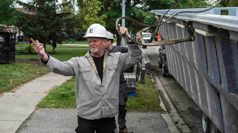 Mike Holmes standing near a dumpster, wearing a hard hat