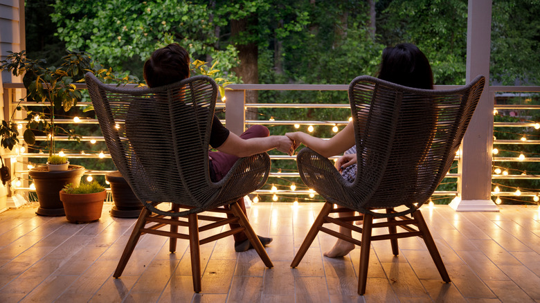 Couple sits in wicker chairs on wood deck with string lights.