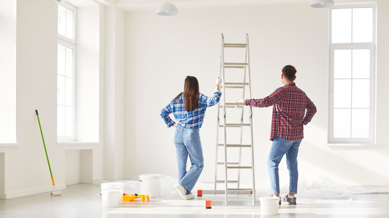 A young couple is looking at a white wall with painting supplies around them