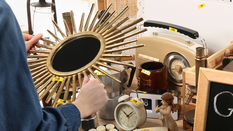 A woman holds an art deco, sunburst style mirror at a thrift store table of antiques.