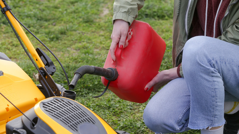 A person kneels to fill up a lawn mower with a red gas can