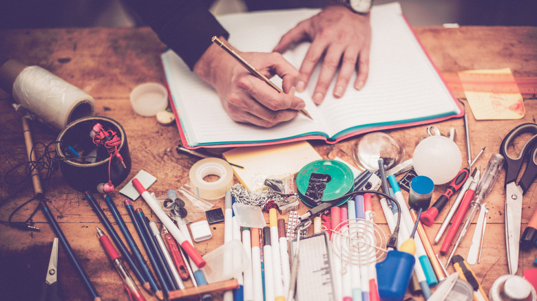 person writing on a table with lots of crafting clutter on it