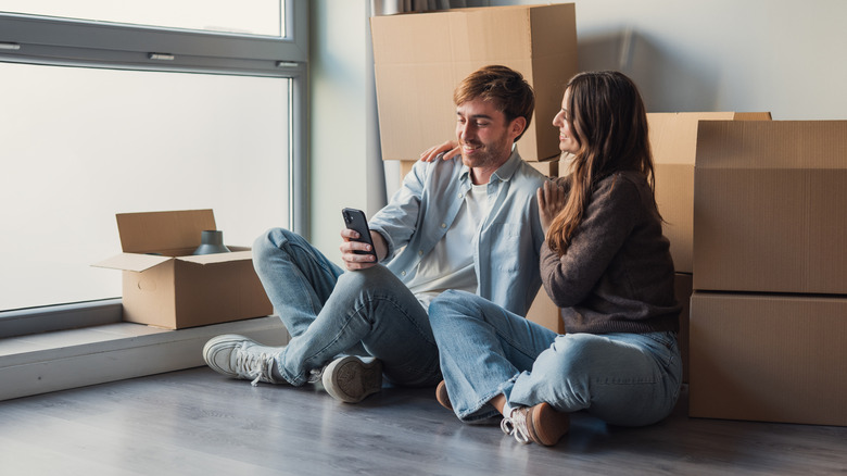 a couple sits with moving boxes on the floor of their home, looking at a phone