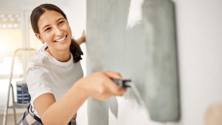 A woman smiles as she rolls on a coat of greenish-gray paint over a white wall.