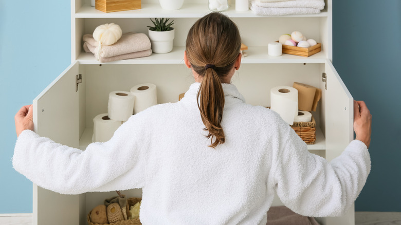 A woman opens up a cluttered bathroom cabinet with stacks of toilet paper