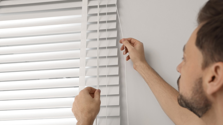 A man is pulling at the cords of traditional blinds covering his windows