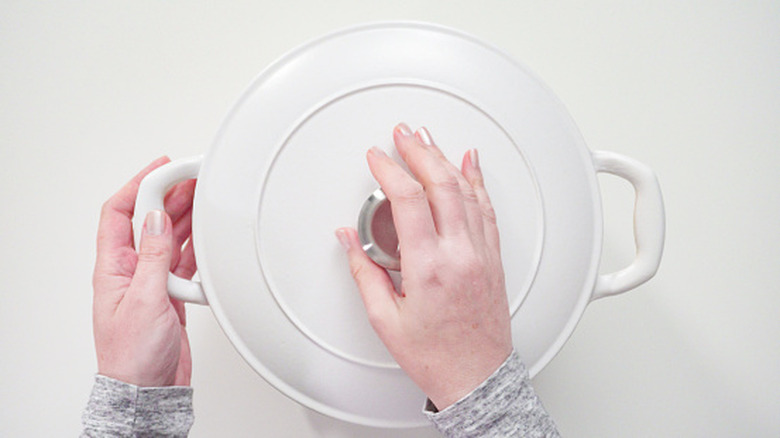 A person with painted nails holds a white pot lid over a white table surface