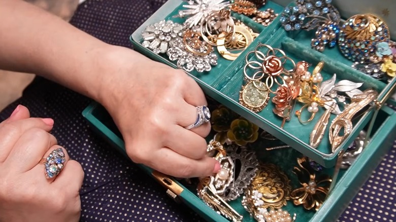 Woman digging through ornate vintage brooch collection in green jewelry box on polka dot tablecloth