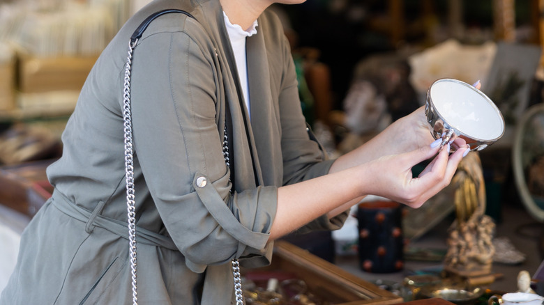 Woman in gray-green coat with manicured hands examining vintage metal teacup at outdoor market
