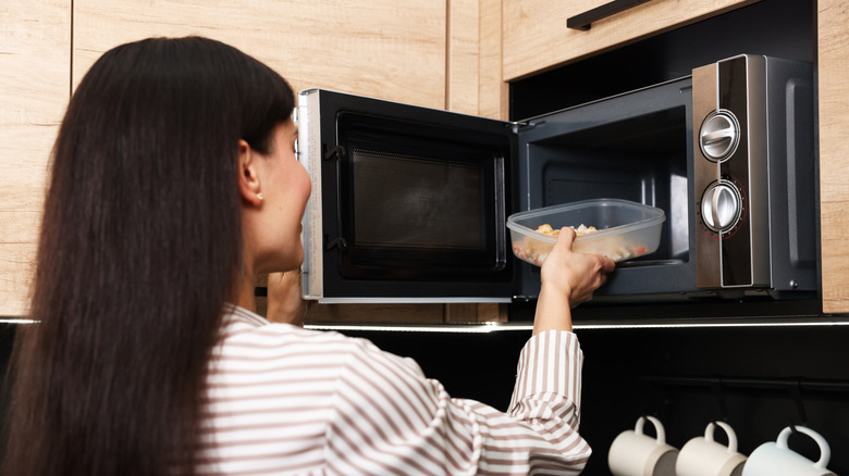 A woman puts a container of leftovers into a microwave to reheat it