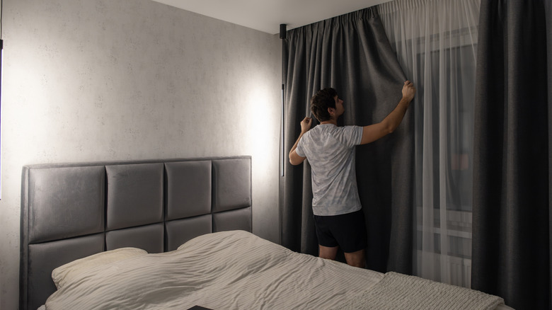 Man closing blackout curtain in modern bedroom
