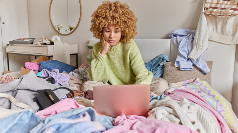 A woman surrounded by clutter on her bed: clothing, blankets, and handbags, plus knickknacks on the table