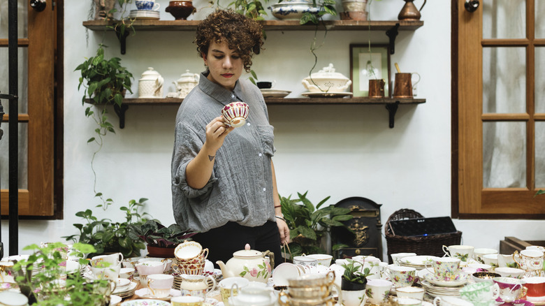 A person holds up an antique teacup in front of a selection of cups, plates, and teapots