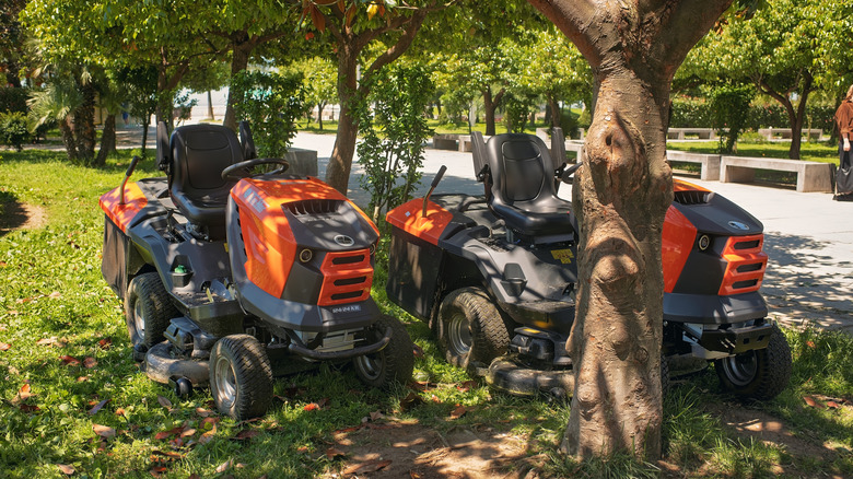 Two riding lawn mowers next to a tree.