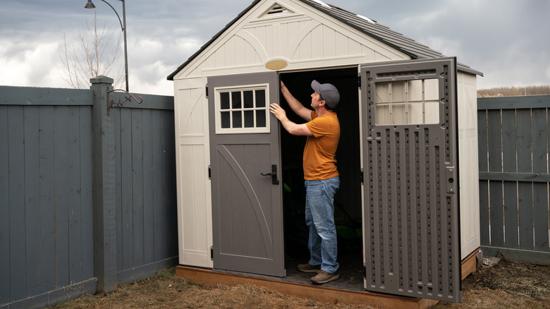 A person wearing jeans, a ball cap, and an orange shirt fixing a door on a utility shed.