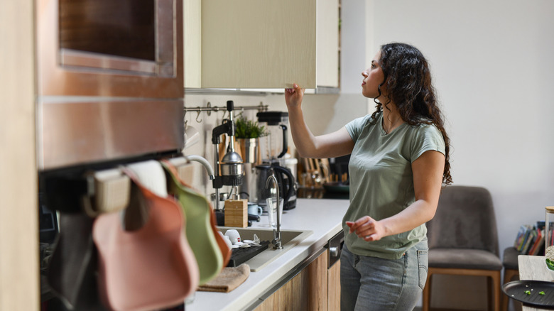 A woman opens a cupboard in a kitchen with cluttered countertops