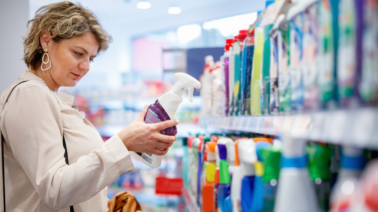 Woman reading product label in store aisle beside shelves of cleaning products.