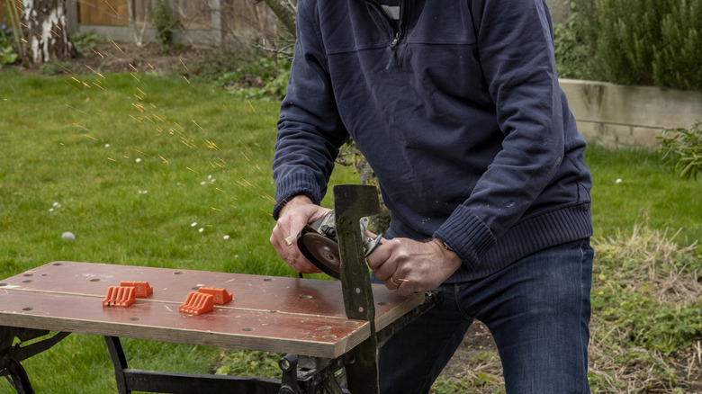 A man is sharpening a dull lawn mower blade with a vice and angle grinder