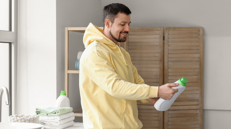 man in yellow shirt holding up white and green laundry detergent bottle with other laundry room elements in the backroom
