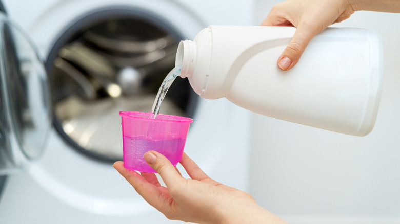 Hands pouring laundry detergent from a bottle into a measuring cup in front of an open washer.
