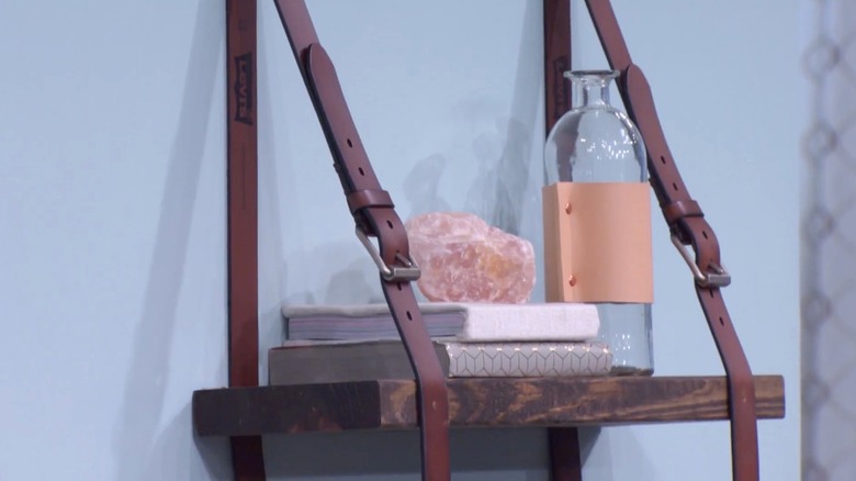 A vintage glass and books rest on a wooden shelf mounted to the wall with two leather belts