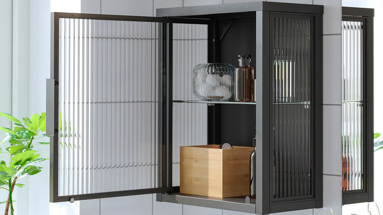 The door of a black MOSSJÖN Wall cabinet with glass door is open to reveal bath products sitting on two shelves
