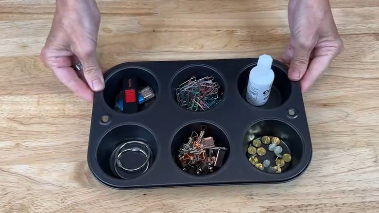A person holds a black vintage muffin tin filled with office supplies