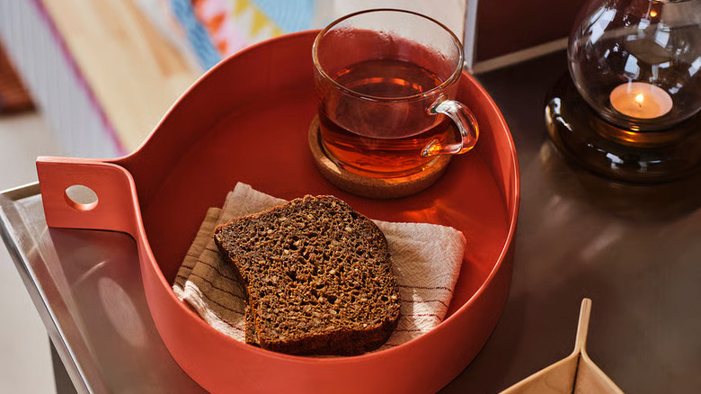 An orange OMMJÄNGE Tray from IKEA filed with a piece of brown bread and a glass mug of tea sits on a wooden table