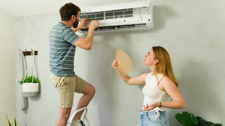 A man is checking the inner parts of an AC while a distressed female fans herself