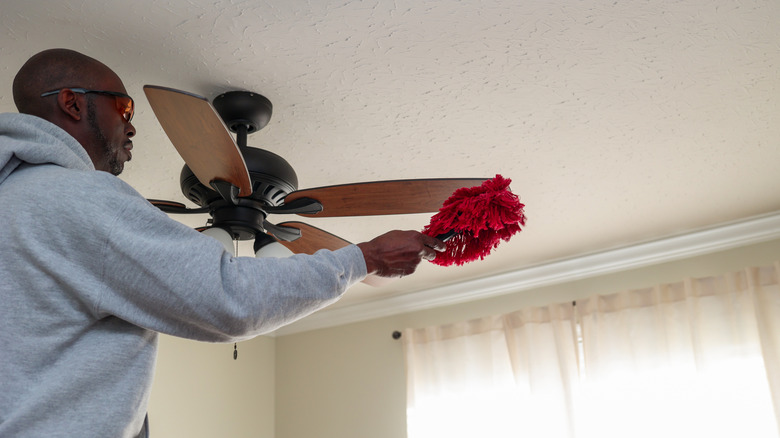 A man is wiping the blades of a ceiling fan with a duster