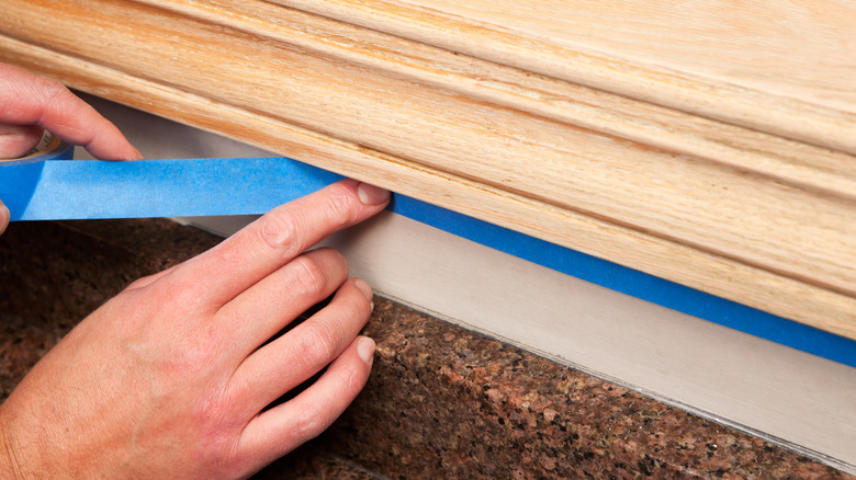 A hand applying blue painter's tape to a surface below wood and above a surface